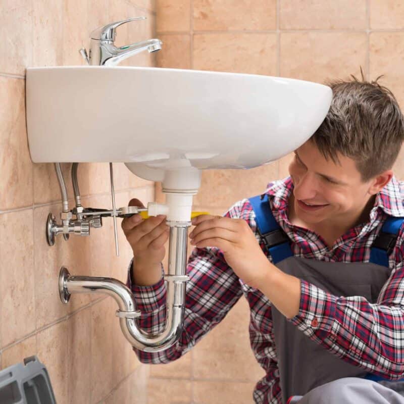 A plumber wearing a plaid shirt and overalls is kneeling on the floor in a tiled bathroom while working on the pipes under a wall-mounted sink. As he uses a wrench to tighten or loosen a pipe fitting, his toolbox sits beside him; he's carefully explaining how to turn off the water before any repairs.