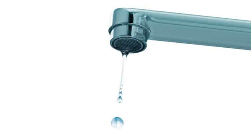 A close-up image of a metallic faucet, possibly installed by a plumber near me, shows a single droplet of water falling from the tap. The background is white, emphasizing the clear and focused water droplet in mid-air.