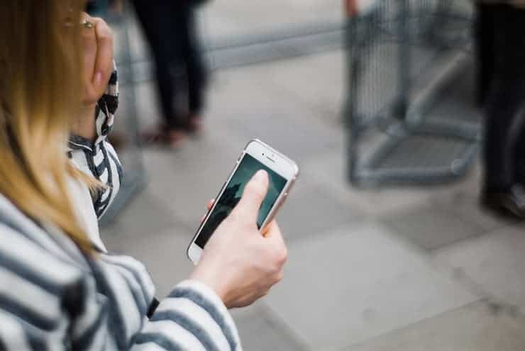 A person with long hair, wearing a striped jacket, holds a smartphone in one hand and touches the screen with the other while standing on a paved walkway. Blurred figures and a metal fence are visible in the background as they perhaps search for a plumber near me.