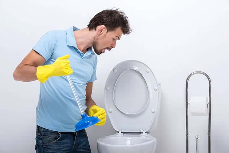 A man wearing a light blue polo shirt and yellow rubber gloves is holding a plunger and looking inside a toilet. He appears to be preparing to unclog it, clearly focused on some bathroom plumbing. A toilet paper holder is visible next to the toilet, and the background is plain white.