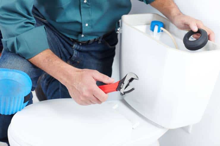 A person wearing a green shirt and blue knee pads is working on bathroom plumbing, using a wrench to fix the inside of a white toilet tank. The toilet lid is open, revealing the tank components. The person is focused on adjusting parts inside the tank.