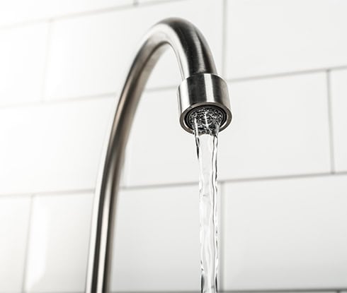 A close-up image of a modern stainless steel kitchen faucet with water jetting from it. The background consists of white subway tiles, adding a clean aesthetic. The faucet, designed for efficient plumbing, features a sleek high arc design.