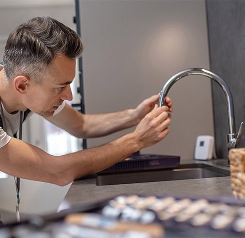 A man with gray hair in a white shirt is focused on fixing a kitchen faucet. He uses a tool while leaning over the sink, ensuring jet cleaning perfection. The background reveals a modern kitchen with a basket and a small electronic device on the counter.