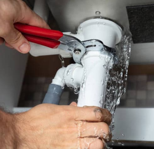 A person using a red-handled pipe wrench to fix a leaking white sink pipe underneath a sink. Water is gushing out from the pipe, highlighting the need for urgent jet cleaning to resolve this plumbing issue.