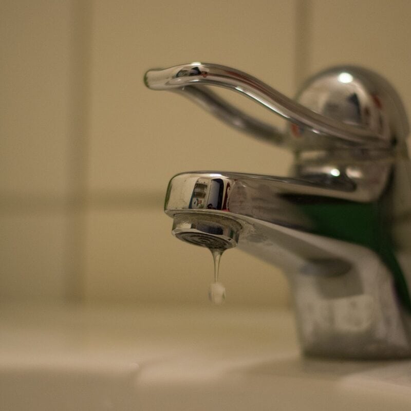 A close-up of a chrome sink faucet with a single handle positioned in a bathroom. The faucet has a drop of water dripping from the spout, indicating a possible leak that may need expert plumbing. The background features white tiled walls.