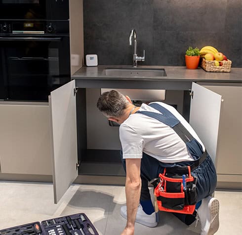A plumber wearing overalls and a tool belt kneels in front of an open cabinet under a kitchen sink, inspecting the plumbing. A toolbox is on the floor nearby, and there are fruits in a tray on the counter. A modern kitchen setup from Reliable Appliance Installation Services is visible in the background.