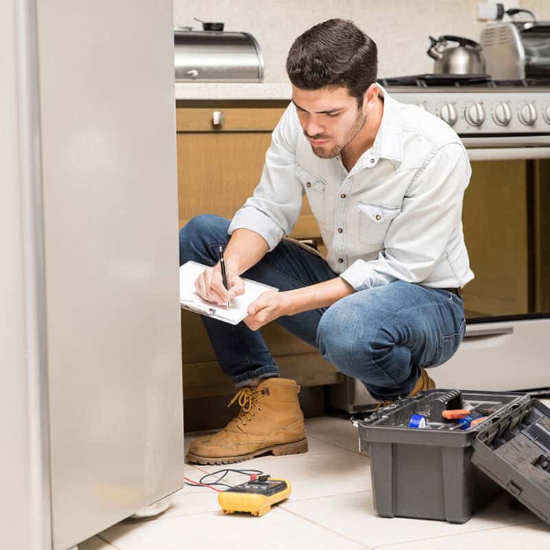 A repair technician kneels on the kitchen floor in front of an open toolbox with various tools, writing on a clipboard. He is examining a refrigerator, with a stove and counter visible in the background. Representing Reliable Appliance Installation Services, he wears work boots, jeans, and a light-colored shirt.