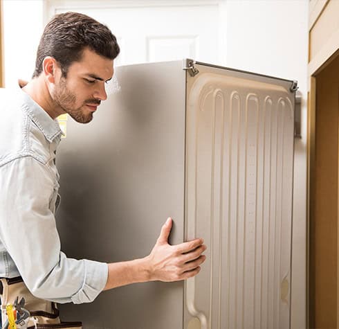 A man with short hair and a beard, wearing a light-colored long-sleeve shirt and a tool belt, is moving a large refrigerator between two cabinets in a kitchen setting. Trust our Reliable Appliance Installation Services for seamless appliance setup every time.