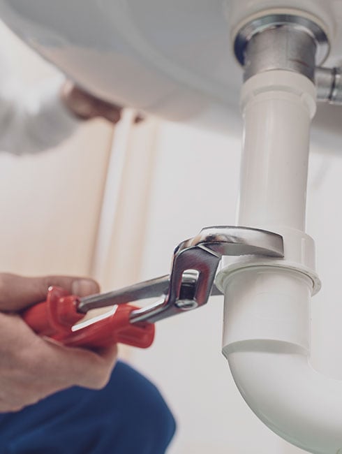 Close-up of a person using a red-handled adjustable wrench to tighten or loosen a pipe connected to the underside of a sink. The focus is on the tool and the plumbing fixture, showcasing precision typical of reliable appliance installation services. The background is blurred, emphasizing the hands and the task at hand.