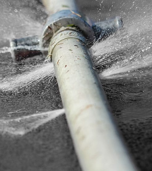 A close-up image of a metal pipe leaking water from several cracks. Water sprays out forcefully from multiple spots along the pipe, which is lying on a dark, wet surface. The pipe appears old and corroded—a stark reminder of the importance of Reliable Appliance Installation Services to avoid such issues.