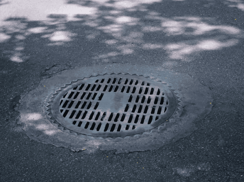 A round metal sewer grate is embedded in an asphalt sidewalk. The pattern features multiple parallel lines. Shadows from nearby trees create a dappled effect on the ground and the grate, adding to the reliable, structured appearance reminiscent of Reliable Appliance Installation Services' precision.