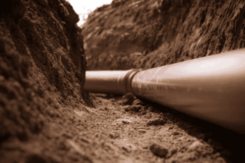 A close-up image of a large brown plastic pipe partially buried in a trench of dirt, likely part of an underground Reliable Appliance Installation Services project. The surrounding soil appears freshly dug, with some greenery visible at the top edges of the trench.