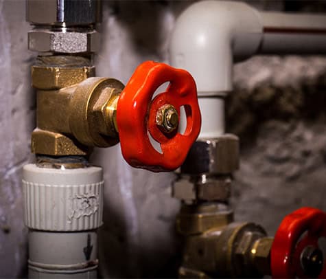 Close-up of a plumbing system featuring metal pipes and fittings. A prominent red valve handle is in the center, used to control the flow of water or gas. The surrounding pipes are connected at right angles, appearing robust and industrial—perfect candidates for thorough plumbing pipes inspection services.