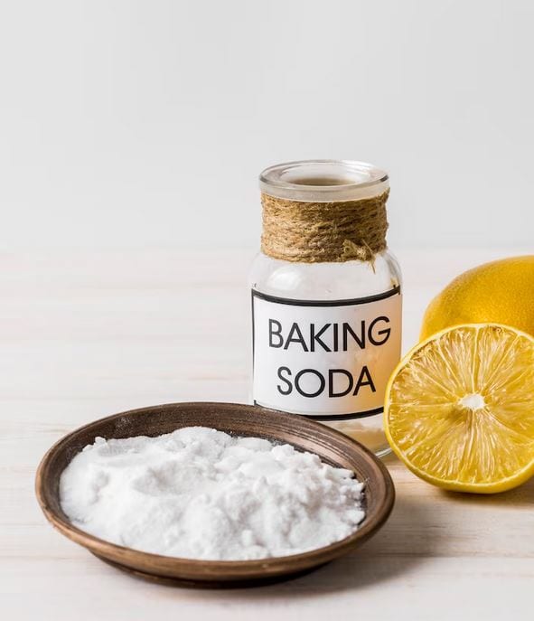 A jar labeled "Baking Soda" with a rope-wrapped neck sits on a light wooden surface. In front is a round wooden dish filled with baking soda. Half of a lemon, with its interior facing up, is beside the jar. Need a reputable plumber? The background is plain white.