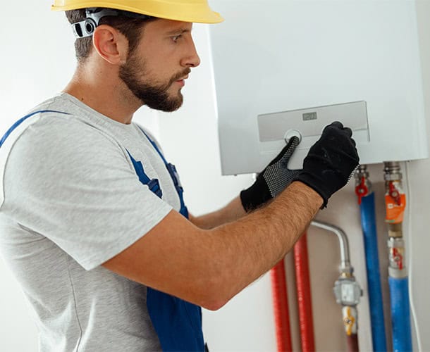 A man wearing a yellow hard hat, gray t-shirt, blue overalls, and black gloves is adjusting a control knob on a wall-mounted device with pipes. The device appears to be part of an HVAC or plumbing system. This plumber in Earlwood works against a white, plain background.