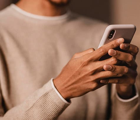 A person wearing a beige sweater is holding and using a smartphone with both hands. Only the lower part of their face and their hands are visible. The background is out of focus, making the person and their phone the main subject of the image. Need a reputable plumber?