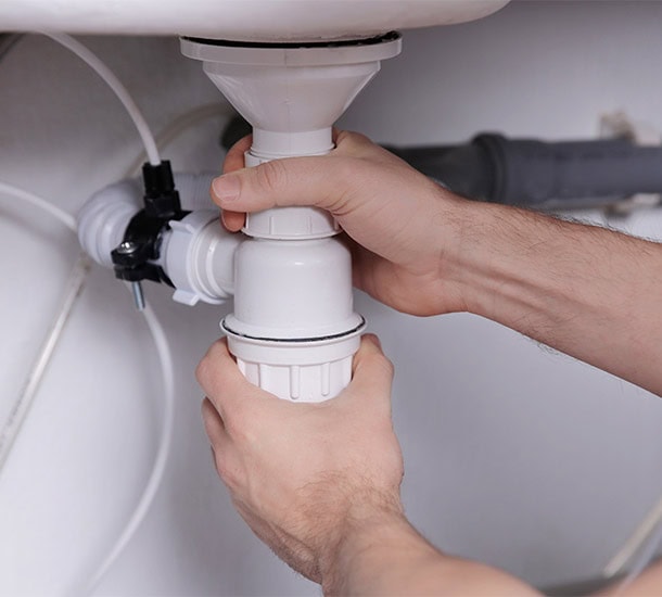 Two hands are fixing a white plastic pipe under a sink. The plumber from Penrith is tightening a coupling joint to secure the piping. Various other pipes and tubing are visible in the background. The scene suggests a plumbing repair or maintenance task.