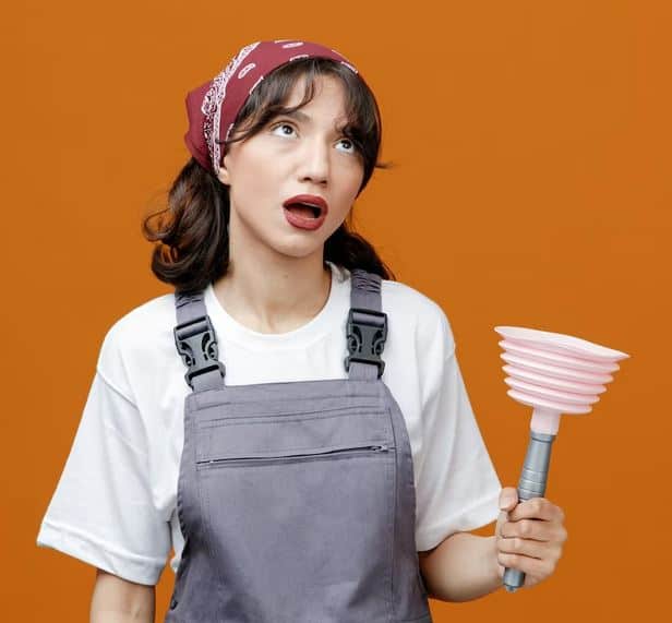 A woman wearing a red bandana, white shirt, and gray overalls holds a pink plunger. With an exasperated expression on her face, she stands in front of an orange background. Need a reputable plumber?