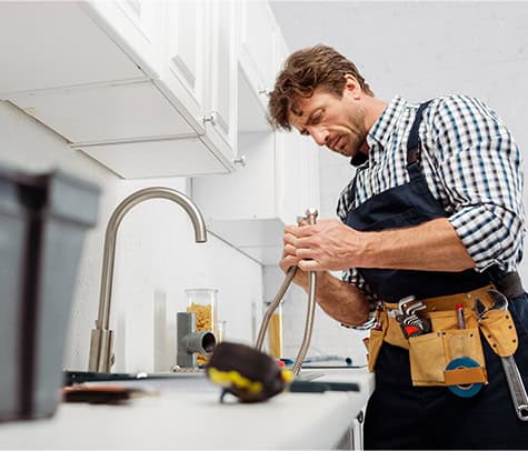 Need a reputable plumber? A man with a tool belt works on plumbing under a kitchen sink. He is wearing a plaid shirt and an apron, and is focused on fixing the faucet. Tools and supplies are visible on the countertop and in a nearby container.
