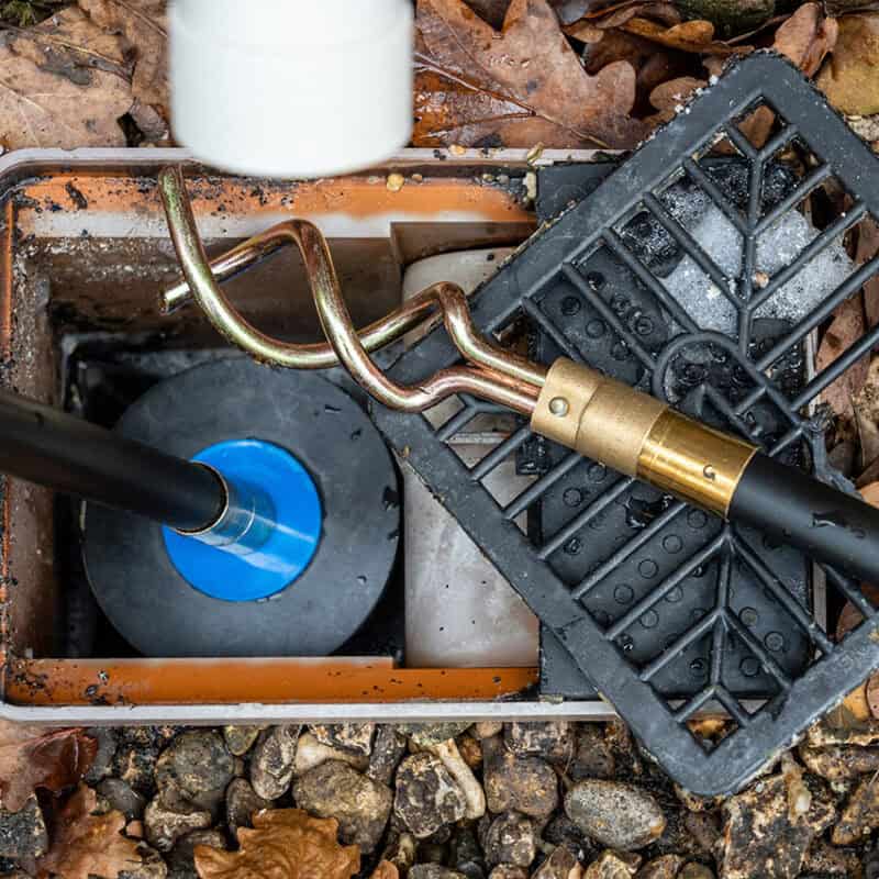 A close-up shot of an open utility box set in the ground among scattered leaves and small stones. The box contains plumbing or electrical components, including a coiled tool and two pipes, one with a blue coupling, useful for addressing issues like "What to Do If Your Sewer Line Is Clogged." The black lid is partially removed and resting nearby.
