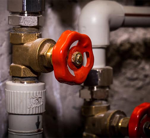 A close-up of a plumbing system features a red valve handle connected to a metallic pipe. The background shows additional pipes and fittings, illustrating part of a larger plumbing network. The scene is well-lit, highlighting common plumbing issues homeowners face with such intricate setups.
