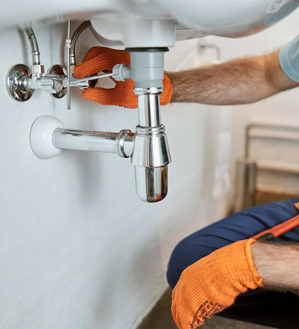 A plumber from Ryde, wearing bright orange gloves, is working under a sink, adjusting plumbing connections. The person uses tools to tighten fittings on the pipes, which are visible against a white wall. Only the plumber's arms and part of the sink and piping are in view.