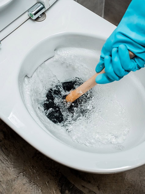 A person wearing blue rubber gloves uses a wooden stick to unclog a toilet. Water is visibly swirling around in the toilet bowl. The close-up image focuses on the hand, the stick, and the toilet bowl, illustrating an effort that makes you wonder: "Can I Repair a Broken Toilet or Should I Replace It?