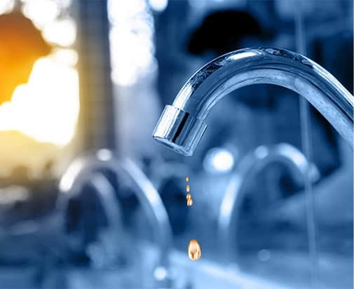 Close-up of a shiny silver faucet with a single water droplet falling from the spout, symbolizing common plumbing issues homeowners face. The background is blurred, showing the outlines of other faucets and a warm light source on the left side, creating a serene and reflective atmosphere.