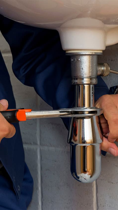 A close-up view of a plumber using an orange-handled wrench to tighten the connection on a chrome P-trap pipe under a white sink. The plumber, wearing a dark blue uniform, works meticulously amidst the light grey tiled wall backdrop. Such attention to detail answers how can I improve water pressure in my home.