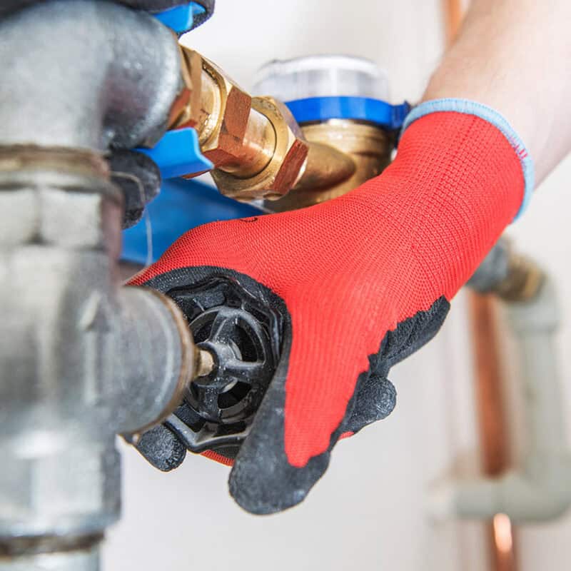 A person wearing red gloves is adjusting a black valve on a metallic pipe system, showcasing one of the common plumbing issues homeowners face. The image focuses on the hands and the valve, with various pipe fittings visible around.