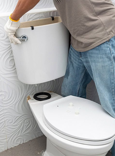 A person wearing gloves and a gray t-shirt is seen installing or repairing the tank of a white toilet in a bathroom with white, embossed patterned walls. The lid of the toilet tank is removed, and the person is adjusting its position, pondering, "Can I repair a broken toilet or should I replace it?
