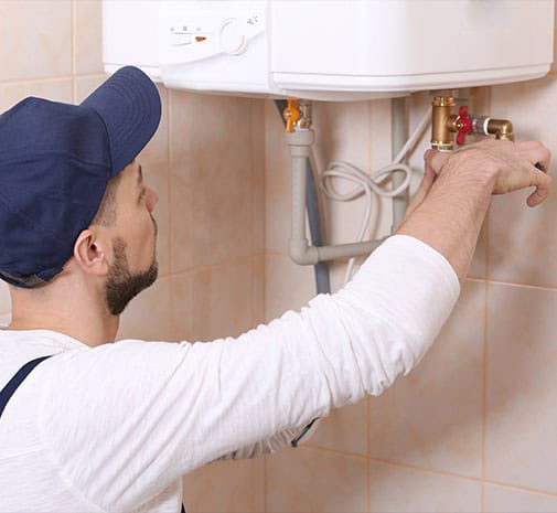 A person wearing a navy blue cap and a white shirt is working on common plumbing issues homeowners face beneath a water heater mounted on a tiled wall. They appear to be adjusting or fixing the pipes attached to the heater.