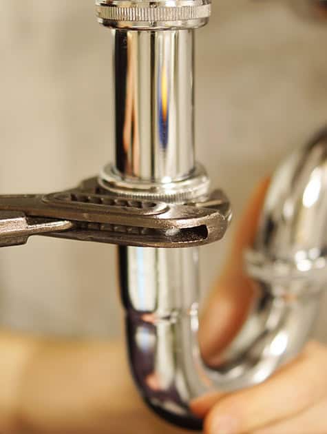 A close-up photograph of a person's hand using a metallic wrench to tighten or loosen a shiny chrome pipe beneath a sink, focusing on the threading of the pipe. The background is blurred, emphasizing the plumbing task—ever wondered how much do plumbers charge now in Australia?