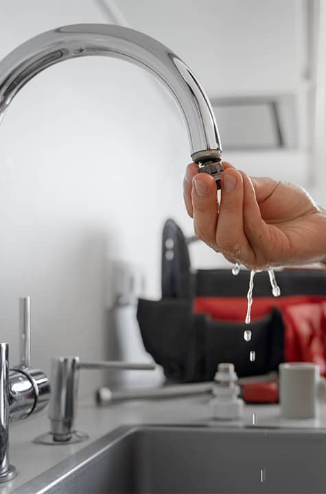 A hand is replacing a faucet aerator in a kitchen sink. Water droplets are falling from the faucet while a toolbox is visible in the background, making you wonder, How Much Do Plumbers Charge Now in Australia?
