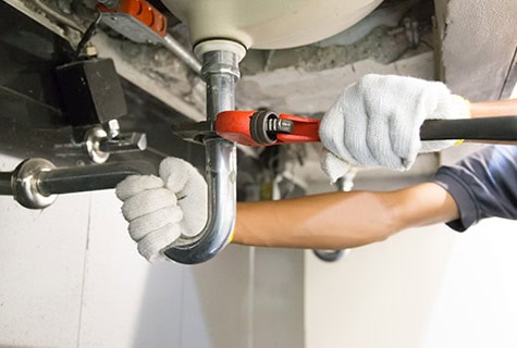 A person wearing gloves uses a pipe wrench to tighten the curved pipe (P-trap) under a sink. The surrounding area shows other plumbing components and fixtures, making one wonder: How much do plumbers charge now in Australia?