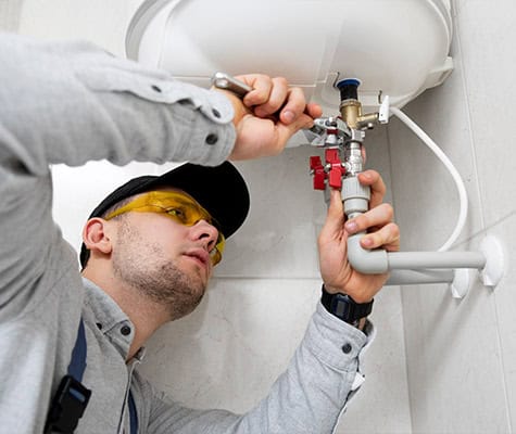 A plumber wearing a cap and protective yellow glasses is using a wrench to fix pipes connected to a water heater. Positioned under the unit in a bathroom setting, he focuses intently on his work, perhaps pondering over the question: How Much Do Plumbers Charge Now in Australia?