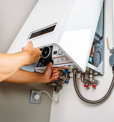 A person is installing or repairing a gas water heater, specifically an instant hot water system. They are shown adjusting parts inside the open front cover. The white wall-mounted unit has visible pipes and connectors beneath it, indicating its plumbing connections.