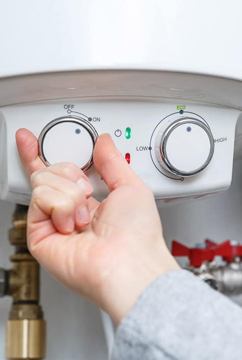 A hand is adjusting the temperature dial on a white water heater. The panel features a power switch with indicators for on and off, two dials labeled "Low" and "High," and an "Eco" mode button. Plumbing components, including the water mains, are visible in the background.