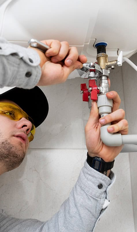 A person wearing a gray shirt, black cap, and yellow safety glasses uses a wrench to tighten a pipe fitting on a white plumbing fixture. The person is focused and working in a bathroom setting with water mains visible near their hands.
