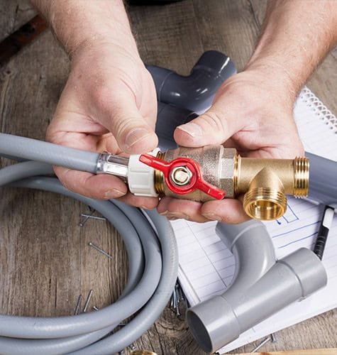 An emergency plumber works on a plumbing project, holding a brass pipe fitting with a red valve. Gray pipes and hoses surround the workspace, along with a notebook and pen in the background on a wooden surface. Various screws are scattered around.