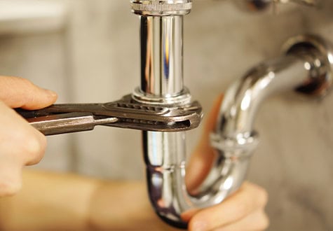 A close-up image of a person using a pipe wrench to tighten or loosen a shiny metal pipe under a sink. The focused area shows the hands of emergency plumbers gripping the wrench and the pipe, with a blurry background.