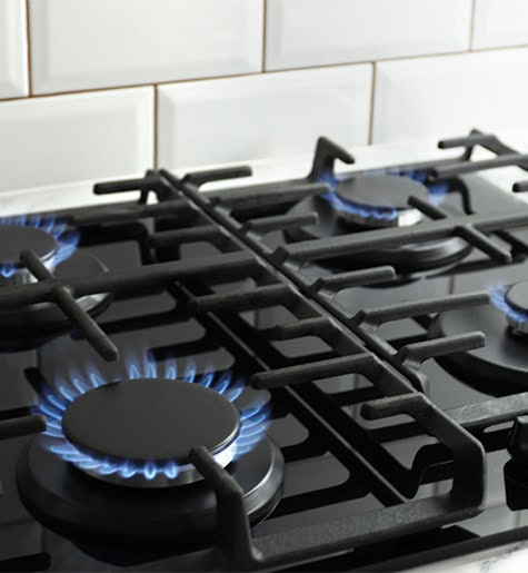 A close-up view of a stainless steel gas stove with four burners turned on, emitting blue flames. The stovetop features black iron grates and is against a white tiled backsplash, ensuring no gas appliance issues as it operates flawlessly.