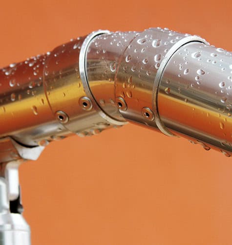Close-up of a metal pipe with joints, covered in water droplets. The shiny pipe's reflective surfaces highlight potential gas appliance issues. The background is a warm orange color.