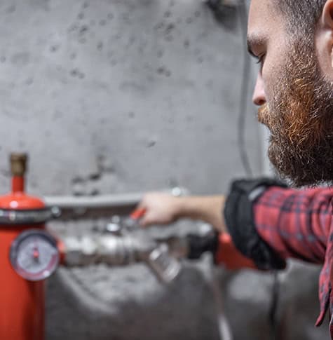A bearded person in a plaid shirt is adjusting a valve on a red pipe, likely addressing gas appliance issues. They are focusing intently on their work in an industrial setting with some gauges and unfocused background elements.