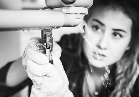 A person wearing gloves uses a wrench to fix a leaking pipe, possibly addressing broader gas appliance issues. Water is seen dripping from the pipe. The person's face is focused on the task at hand. The image is in black and white.