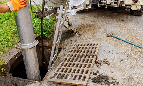 A worker in orange gloves is conducting plumbing maintenance, using equipment to clean a sewer drain. The metal grate from the drain is lifted and placed to the side, attached with a chain. There is a ladder leading down into the sewer, and a blue tool is on the ground nearby.