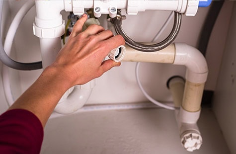 A person is performing plumbing maintenance under a sink, focusing on a pipe connection. The individual is holding a pipe joint with one hand and appears to be adjusting or replacing it. Various pipes and connectors are visible in the compact space under the sink.