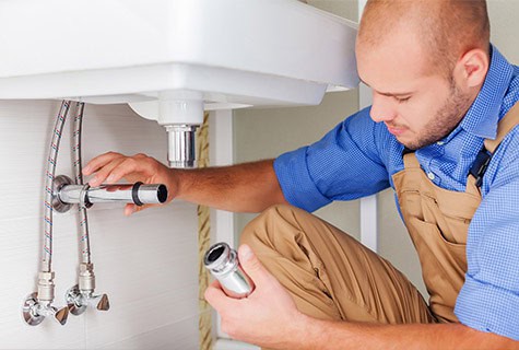 A plumber wearing a blue shirt and beige overalls is crouched under a white sink. He is holding a pipe in one hand and inspecting the plumbing connections with the other, ensuring proper plumbing maintenance. Various pipes and valves are visible next to him.