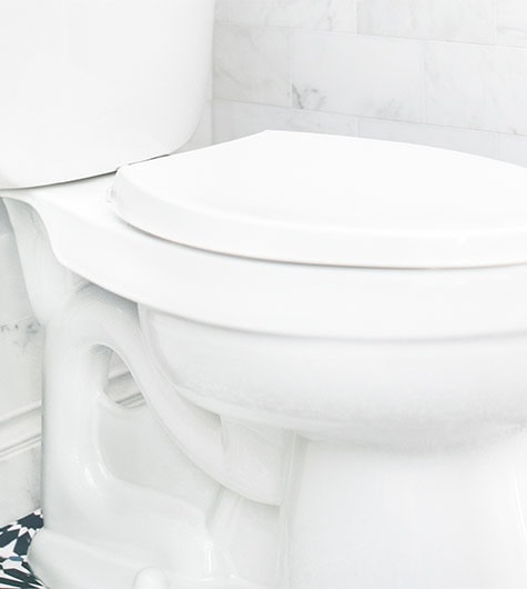 A close-up image of a white toilet with the lid closed. The toilet, part of a green plumbing initiative, is installed against a tiled wall with a marble design. The flooring features a black and white geometric pattern. The image emphasizes the clean and modern bathroom setting.