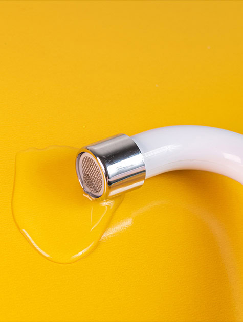 A white hose with a metallic nozzle, part of a green plumbing setup, is leaking water onto a bright yellow surface.